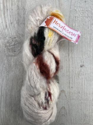 Ein heller Strang Suri Alpaka mit farbigen Sprenkeln in Schwarz, Gelb, Rotbraun und Pink, handgefärbt, liegend auf einem Holzbrett.